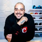 Berner smiling and leaning on a counter inside a Cookies retail store, wearing a black sweatshirt with the Cookies logo and a tattoo of the word “Cookies” on his hand. Shelves of branded hats are visible in the background.