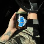 Person holding a dark glass jar with a Cookie Monster sticker labeled “Cookie Dough,” sitting in a car with sunlight highlighting tattoos and camouflage clothing.